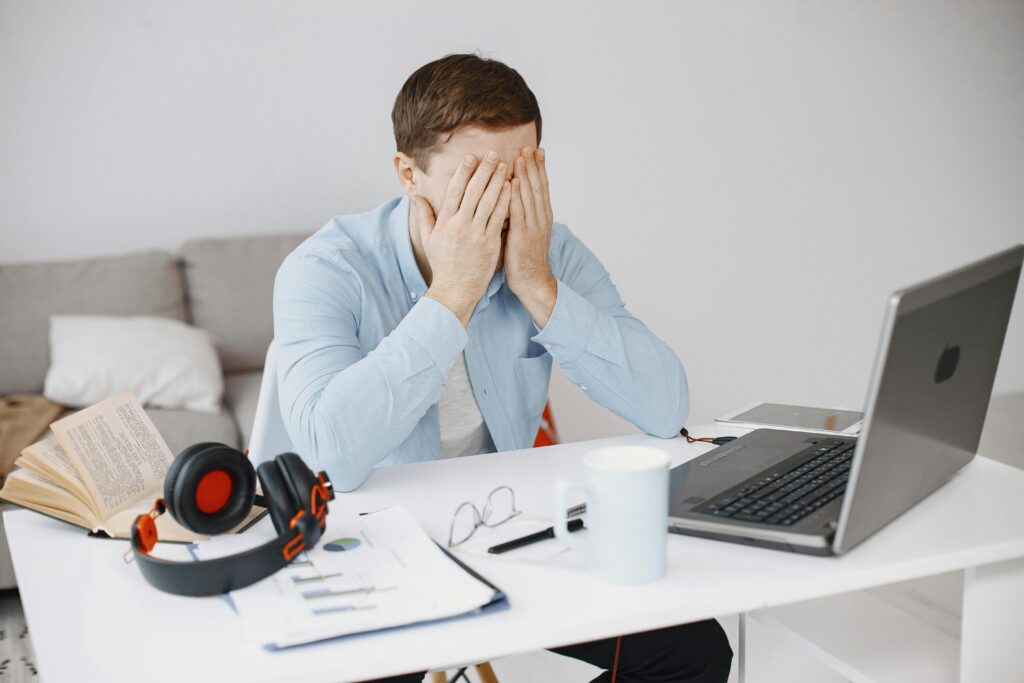 A person sitting at a desk with their face covered by their hands, appearing stressed or overwhelmed, with a laptop, papers, and headphones nearby.
