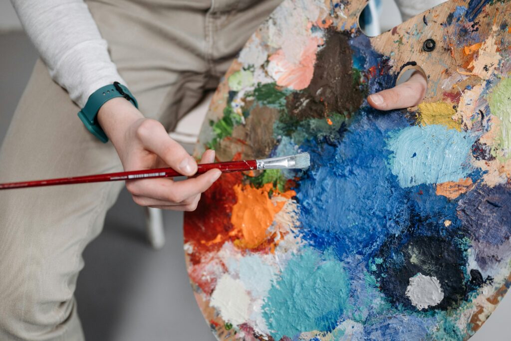 Close-up of an artist holding a paintbrush over a colorful paint palette filled with vibrant mixed colors