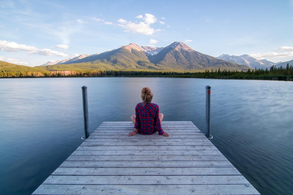 A person sits quietly at the end of a wooden dock overlooking a calm lake, with mountains rising in the background under a clear blue sky, creating a peaceful and serene scene of silence and reflection.