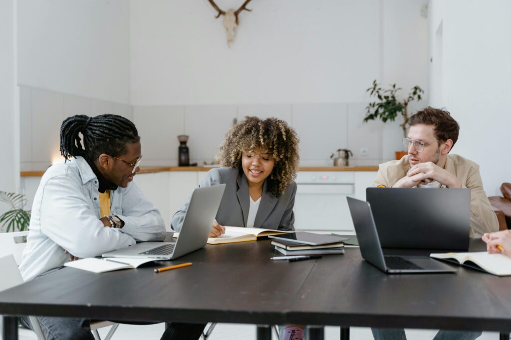 Three professionals sit around a conference table in a modern office, collaborating on laptops and reviewing documents during a meeting.